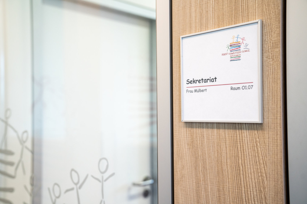 Das Hinweisschild für das Sekretariat neben einer Glastüre mit dem Logo der Schule Das Hinweisschild für das Sekretariat neben einer Glastüre mit dem Logo der Schule