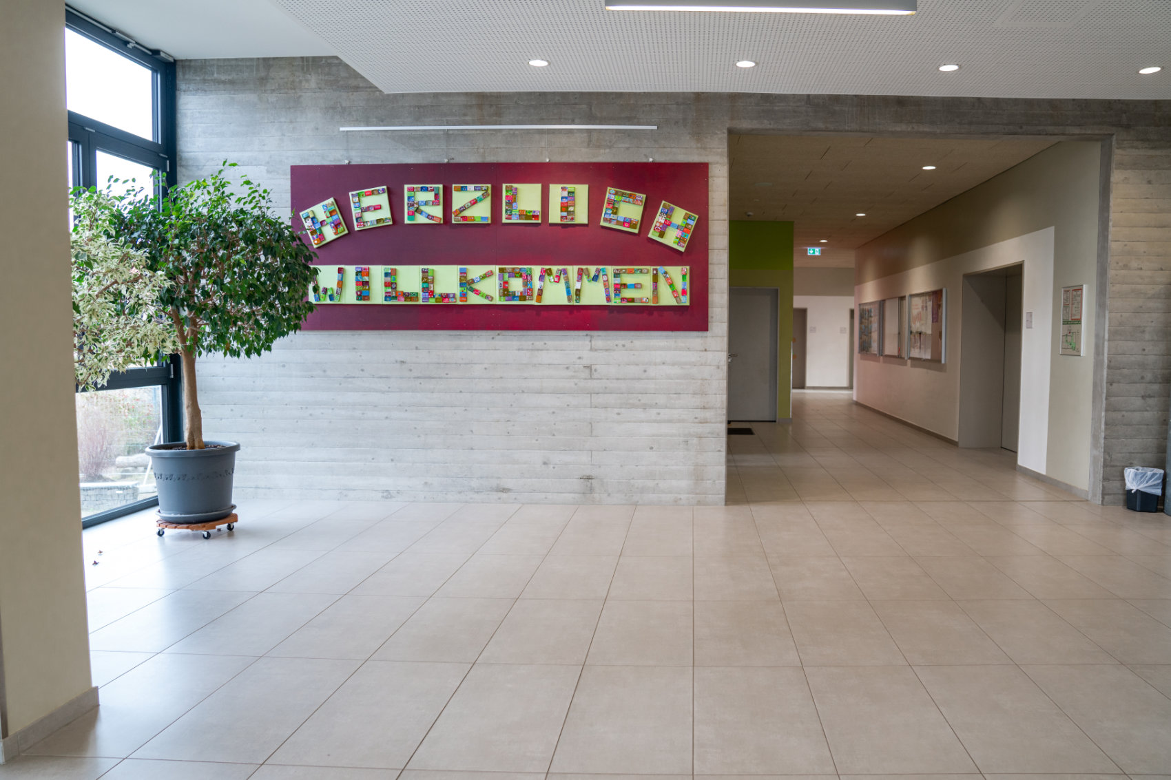 Eingangsbereich mit Ficus Benjamina links vor der Fensterwand und großer Kollage mit Herzlich Willkommen aus bunten Klötzchen an der Wand Eingangsbereich mit Ficus Benjamina links vor der Fensterwand und großer Kollage mit Herzlich Willkommen aus bunten Klötzchen an der Wand