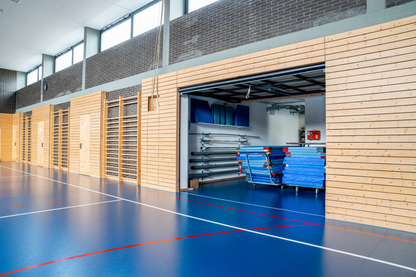 Sporthalle mit blauem Boden und holzverkleideten Wänden 1 Wandsegment aufgeklappt mit Sicht auf 2 Turnmattenstapel Sporthalle mit blauem Boden und holzverkleideten Wänden 1 Wandsegment aufgeklappt mit Sicht auf 2 Turnmattenstapel